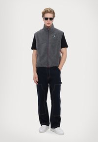 Gray fleece vest with a high collar and front zipper. Worn over a black t-shirt and paired with navy pants. White sneakers complete the outfit.