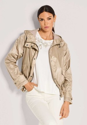 Frau mit dunklen Haaren, die eine beige Jacke mit Reißverschluss über einem weißen Top mit Blumenstickerei und weißen Hosen trägt, steht vor einem schlichten Hintergrund.