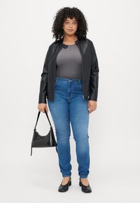 Juodas dirbtinės odos švarkas, pilkas marškinėlis, mėlynos džinsinės kelnės, juodi bateliai plokščiomis padais ir mažas juodas rankinukas. Garbanoti plaukai, stovintys priešais baltą foną.