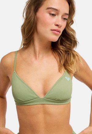 Kobieta o falowanych włosach, ubrana w prążkowany, jasnozielony bikini top na cienkich ramiączkach, patrząca w dół i na lewo, na jednolitym tle.