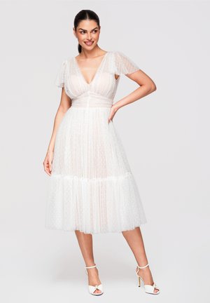 Femme souriante, portant une robe blanche mi-longue avec des manches papillon transparentes et une texture à pois, associée à des sandales blanches à talons hauts.