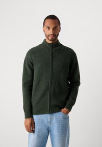 Mann iført en mørkegrønn genser med full glidelås og lyseblå jeans, stående med den ene hånden i lommen mot en ensfarget bakgrunn.