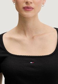 Černý žebrovaný top s hranatým výstřihem a krajkovým lemem, ozdobený malým logem v červené, bílé a modré barvě uprostřed.