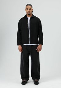 Zwart overhemd over een wit T-shirt, gecombineerd met brede zwarte broek en zwarte schoenen. De outfit heeft een ontspannen pasvorm en een minimalistisch ontwerp.