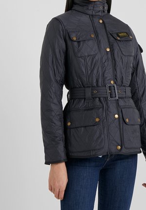 Czarna pikowana kurtka z wysokim kołnierzem, kieszeniami z patkami na guziki, paskiem z klamrą, noszona z niebieskimi dżinsami.