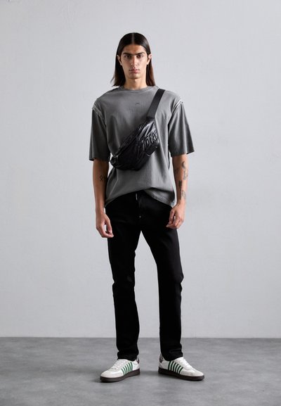Junger Mann mit langen Haaren steht an einer grauen Wand, trägt ein übergroßes dunkelgraues T-Shirt, schwarze Hosen, weiße Sneakers und eine schwarze Umhängetasche.
