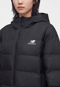 Άτομο που φορά μαύρο φουσκωτό μπουφάν New Balance με κουκούλα και μπροστινό φερμουάρ, εμφανίζοντας την περιοχή του στήθους και το λογότυπο.