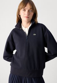 Navy blue zip-up sweatshirt with textured fabric, ribbed hem, and a small green logo on the left chest. Model wears a white collared shirt underneath.