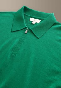 Polo vert en maille avec une fermeture demi-zip et un col plié, affichant une étiquette de marque blanche cousue au niveau de l'encolure.