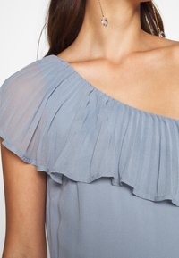 Haut bleu clair avec un plissé unique sur l'épaule, fabriqué en tissu léger et transparent, avec une texture lisse.