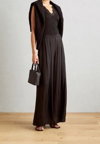 Černý top s žebrovanou texturou a krajkovými doplňky, v kombinaci s volnými černými kalhotami, tmavým kardiganem přehozeným přes ramena a malou černou kabelkou.