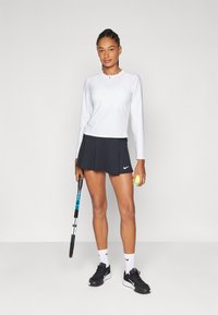 Top atlético branco de manga comprida, saia preta plissada, sapatilhas pretas, segurando uma raquete de ténis e uma bola de ténis amarela. Logótipo da Nike visível.