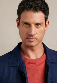 Zbliżenie na mężczyznę w granatowej kurtce i rudym podkoszulku. Włosy są krótkie, ciemne z delikatną falą; ma niebieskie oczy.