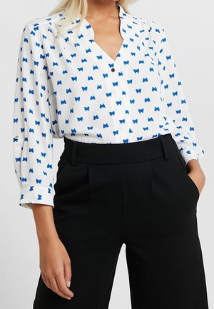 Mujer con blusa blanca con estampado de mariposas azules y pantalones negros de talle alto, una mano en el bolsillo, sobre un fondo liso.