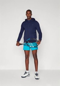 Temno modra jopa z zadrgo, turkizno modre kratke hlače, beli supergi; drži teniški lopar z modro in črno zasnovo. Gladek material, športno prileganje.