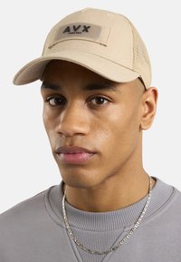 Beżowa czapka baseballowa o zaokrąglonym kształcie, z siateczkowymi panelami i czarną naszywką "AVX" z przodu. Noszona przez osobę w szarej bluzie.