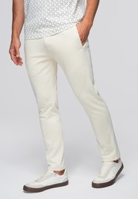 Weiße, schmal geschnittene Sweatpants mit Seitentaschen, aus weichem Stoff. Kombiniert mit weißen Sneakers mit braunen Sohlen und Schnürsenkeln.