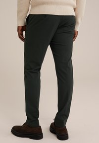 Pantaloni modelați de culoare verde închis, cu o textură netedă, având o croială slim, buzunar la spate și închidere cu nasture, purtați cu pantofi din suede maro.