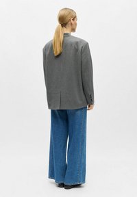 Veste grise surdimensionnée avec épaules rembourrées, fente unique et poignets à trois boutons, associée à un jean en denim bleu à jambes larges. Chaussures plates noires.