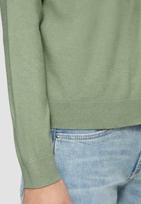 Zielony sweter robiony na drutach z ściągaczami na rękawach i dolnym brzegu, charakteryzujący się gładką teksturą. Noszony z jasnoniebieskimi dżinsami, widocznymi na dole.