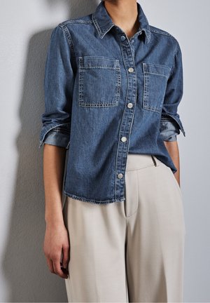 Frau trägt ein blaues Jeanshemd mit Knöpfen und Brusttaschen sowie hoch taillierte beige Hose.
