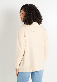Sweat à capuche beige avec une finition texturée, coupe décontractée et ourlet côtelé, associé à un jean bleu clair. Aspect naturel et doux.