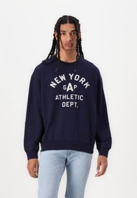 Sudadera azul marino con cuello redondo y hombros caídos. Presenta un texto en blanco: "NUEVA YORK GAP DEPARTAMENTO DE ATLETISMO" en estilo vintage.