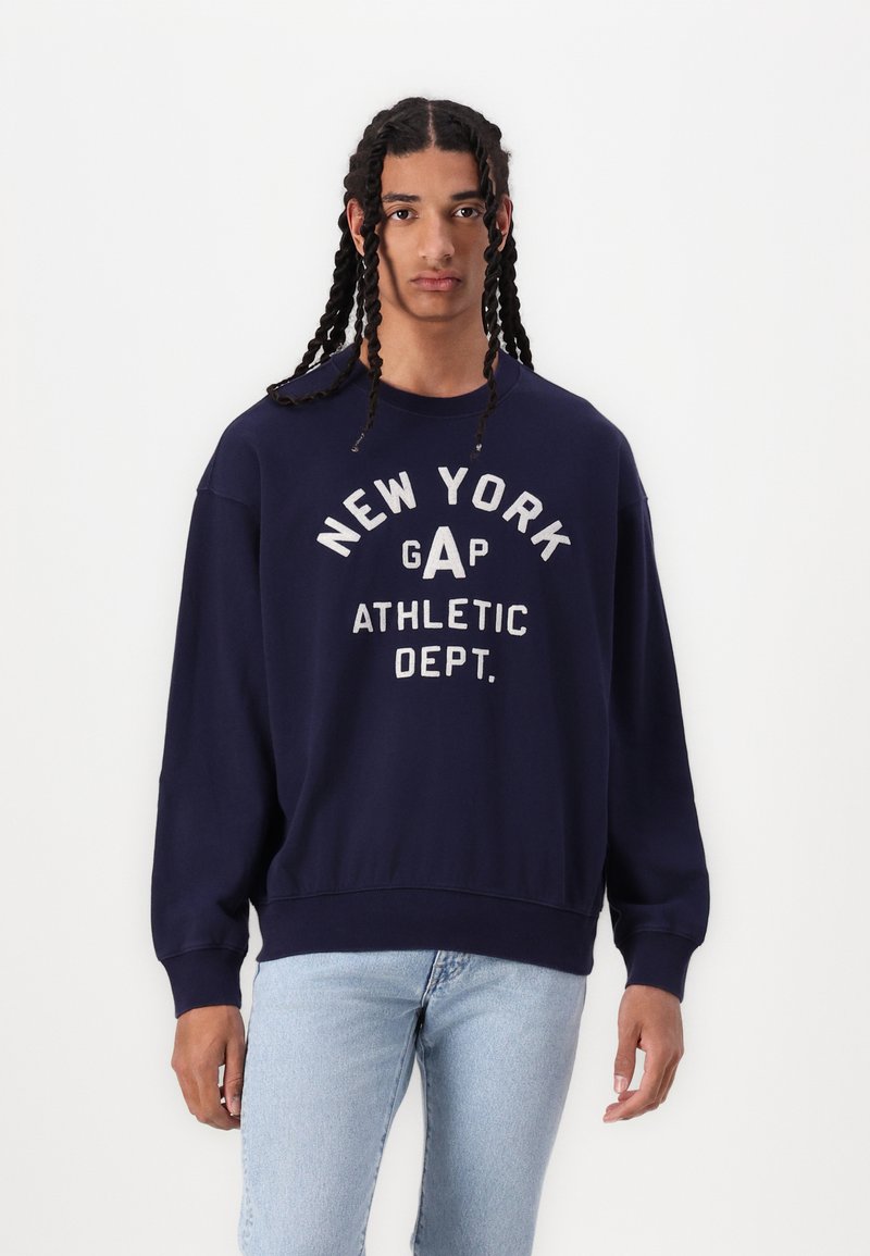 Sudadera azul marino con cuello redondo y hombros caídos. Presenta un texto en blanco: "NUEVA YORK GAP DEPARTAMENTO DE ATLETISMO" en estilo vintage.