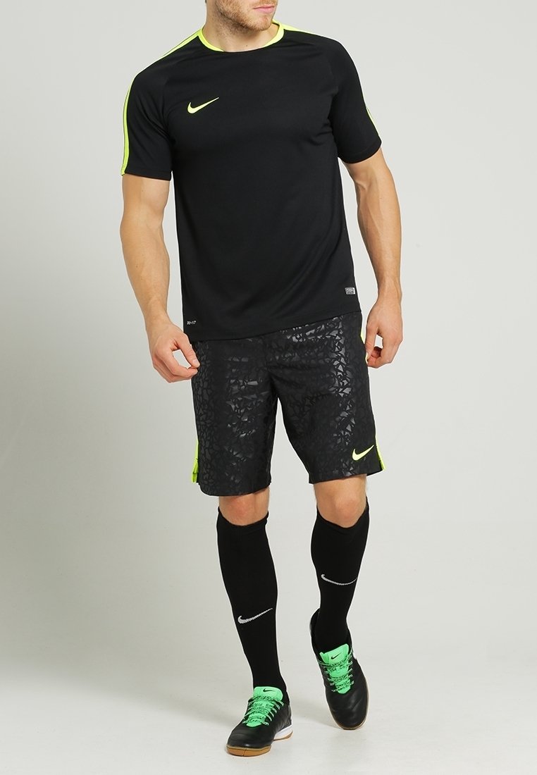 T-shirt de sport Nike noir à manches courtes, avec accents jaune néon. Short noir à motif géométrique. Chaussettes noires et chaussures vertes et noires.