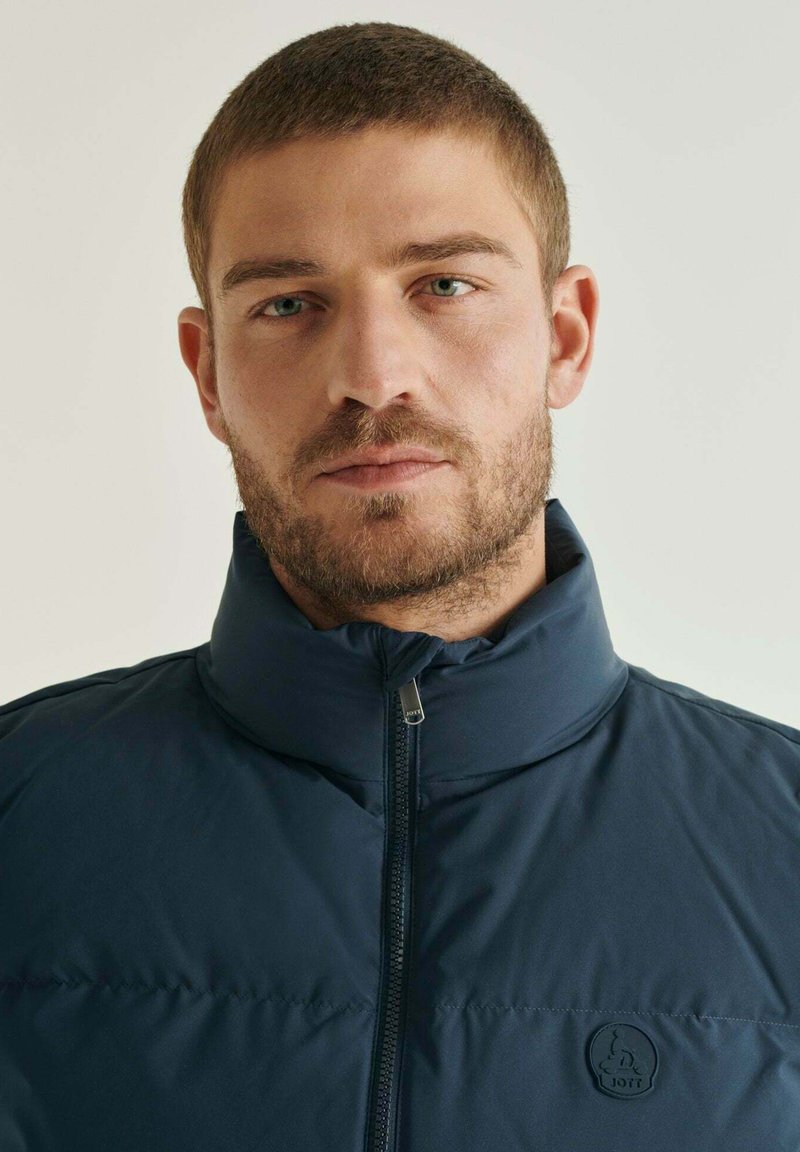 Chaqueta acolchada azul marino con un cuello alto, cremallera completa y un logotipo circular en el lado izquierdo del pecho. Textura suave, diseño ligero.