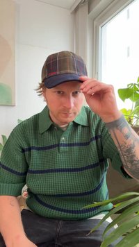 Suéter polo verde em malha com riscas horizontais azuis-marinho, boné de basebol preto com padrão aos quadrados, tatuagem visível no antebraço, planta interior ao fundo.