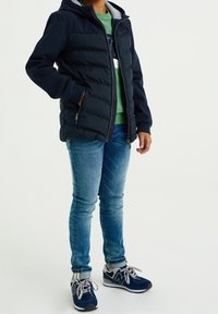 Navy puffer jacket with a hood and gray lining, paired with a green shirt, blue jeans, and navy sneakers. Zipper and pockets visible.