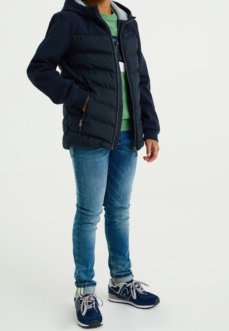 Navy puffer jacket with a hood and gray lining, paired with a green shirt, blue jeans, and navy sneakers. Zipper and pockets visible.
