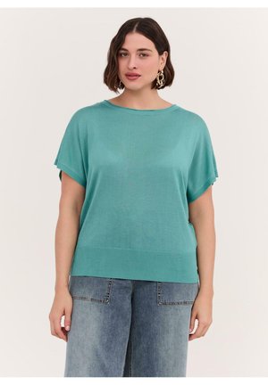 Kobieta z krótkimi ciemnymi włosami, nosząca złote kolczyki-koła, luźną krótka bluzkę w kolorze morskim i jasnoniebieskie dżinsy z szerokimi nogawkami, stojąca na jednolitym tle.