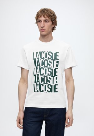 Ung mand med kort krøllet hår iført en hvid T-shirt med mørkegrøn gentaget "LACOSTE" tekst og mørkeblå jeans mod en ensfarvet baggrund.