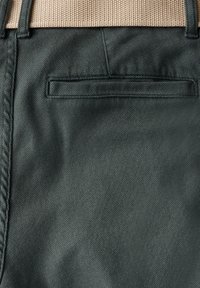 Tela de un verde oscuro con una textura sutil, que presenta una cinturilla de beig tejida y un bolsillo trasero con una abertura oculta.