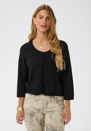 Vrouw met lang golvend haar, gekleed in een zwarte vest met knopen en beige broek met bloemmotief, geaccentueerd met gouden sieraden.