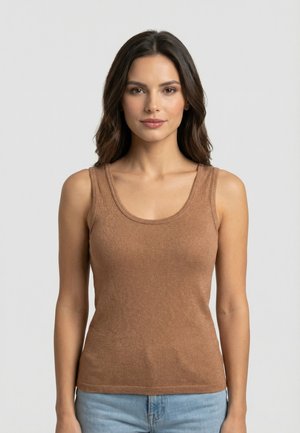 Donna con lunghi capelli scuri, indossa una canotta marrone e jeans azzurri, in piedi davanti a uno sfondo grigio chiaro uniforme.