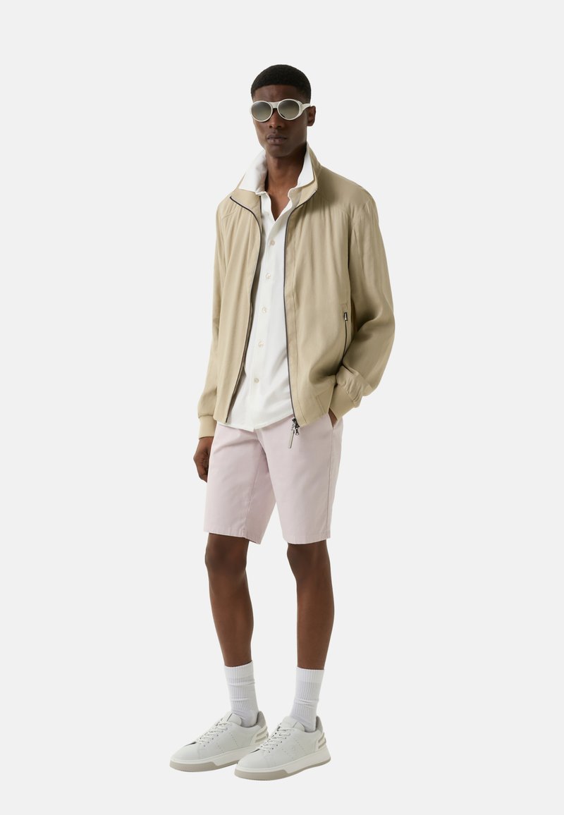 Hellbeige Hoodie, weißes Hemd mit Knöpfen, blassrosa Shorts, weiße Sneakers und ovale Sonnenbrille vor einem weißen Hintergrund.