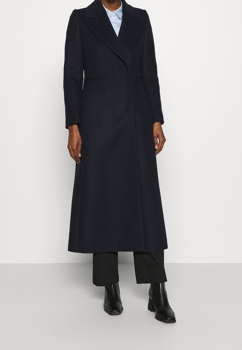 Long navy coat in wool blend, tailored design with notch lapels, side pockets, and a clean silhouette, paired with black heeled boots.