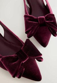 Jedwabiste, burgundowe buty typu slingback z ostrymi noskami i wyraźną kokardą na przodzie. Gładka tekstura z błyszczącym wykończeniem.
