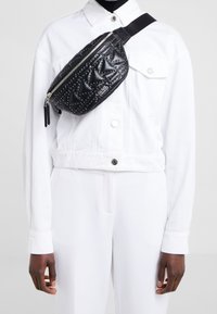 Henkilö, joka käyttää valkoista takkia ja housuja sekä mustaa tikattua olkalaukkua, jossa on pieniä hopeisia prässättyjä helmiä.