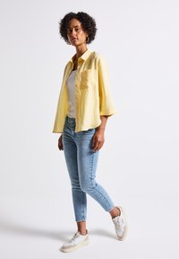 Geel kortemouwen shirt, witte tanktop, lichtblauwe strakke jeans en beige sneakers. Model staat voor een neutrale achtergrond.