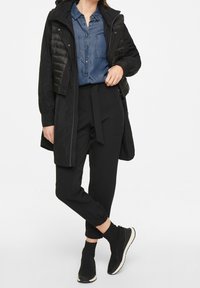 Black puffer jacket layered over a blue denim shirt, paired with black tapered pants. Model wears black ankle boots with white soles.