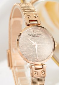 Orologio con cinturino in maglia oro rosa e quadrante argentato texture, caratterizzato da semplici indicatori delle ore e dal logo "Victoria Hyde London."