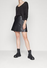 Svart läder A-linje kjol med knapprad, kombinerad med en mörk stickad tröja och svarta ankelstövlar mot en neutral bakgrund.