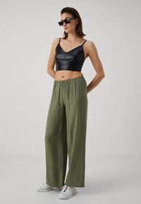 Fekete, sima anyagú cropped topp, zöld, könnyű anyagú, bővülő nadrág, fehér sportcipővel és fekete, négyzet alakú napszemüveggel viselve.