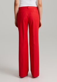 Pantalones ajustados rojos con frente plano, piernas largas y bolsillos traseros. Tejido suave con un acabado nítido, diseñado para una silueta ajustada.