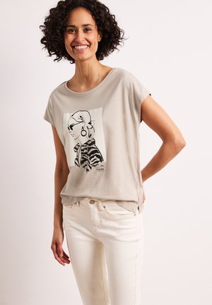 Kvinde med krøllet hår, smilende, iført en beige t-shirt med en sort-hvid skitse af en kvinde med solbriller og hvide bukser.