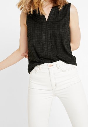 Žena v černé vzorované halence bez rukávů zastrčené do vysoké bílé džíny na jednobarevném pozadí.